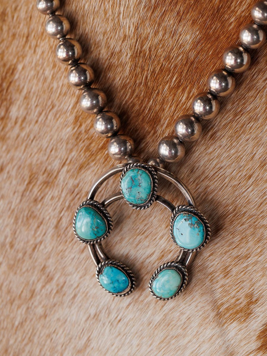 The Pawnee City Necklace by Savannah Sevens western life{&}style is a Native American handmade sterling silver piece with round beads and a five-stone turquoise naja pendant, shown on a brown and white cowhide background.