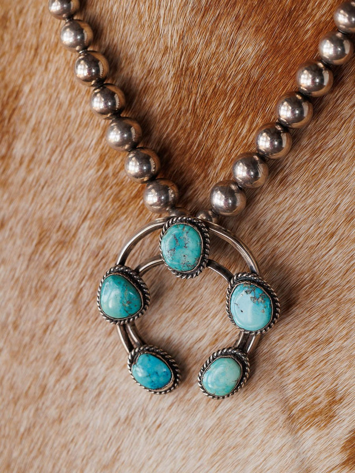 The Pawnee City Necklace by Savannah Sevens western life{&}style is a Native American handmade sterling silver piece with round beads and a five-stone turquoise naja pendant, shown on a brown and white cowhide background.