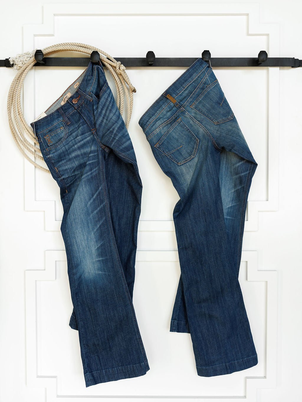 Two pairs of Ariat The Payson Perfect Rise Slim Trouser Jeans in medium dark wash hang by their belt loops on a black wall-mounted rack. A coiled rope hangs to the left against a white wall with decorative trim.