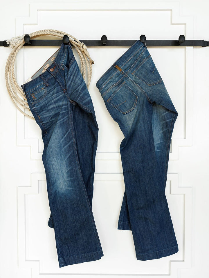 Two pairs of Ariat The Payson Perfect Rise Slim Trouser Jeans in medium dark wash hang by their belt loops on a black wall-mounted rack. A coiled rope hangs to the left against a white wall with decorative trim.