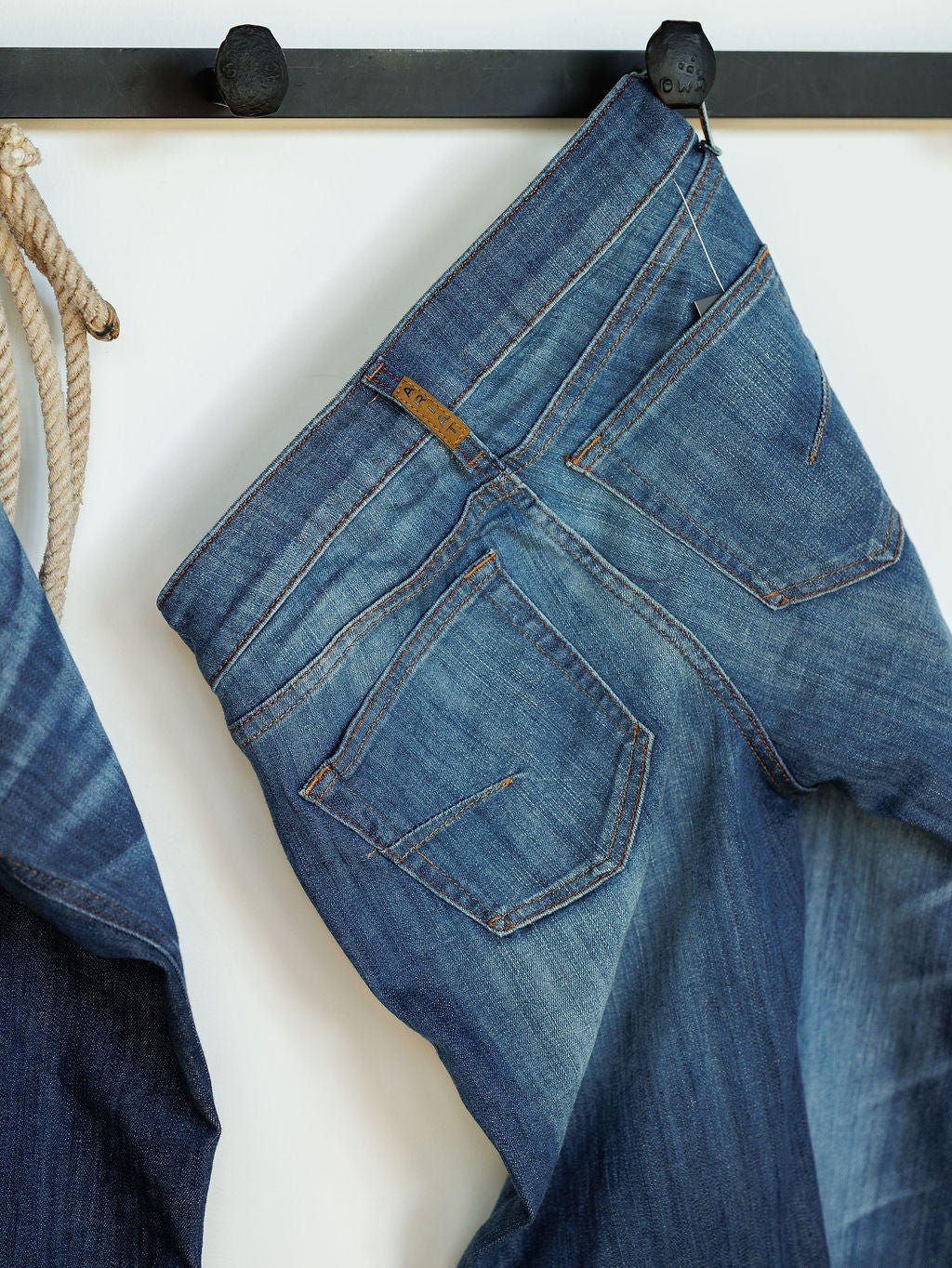 A pair of Ariat The Payson Perfect Rise Slim Trouser Jeans in medium dark wash hang by a belt loop on a black wall hook, showing back pockets and faded details, with a coiled rope beside them.