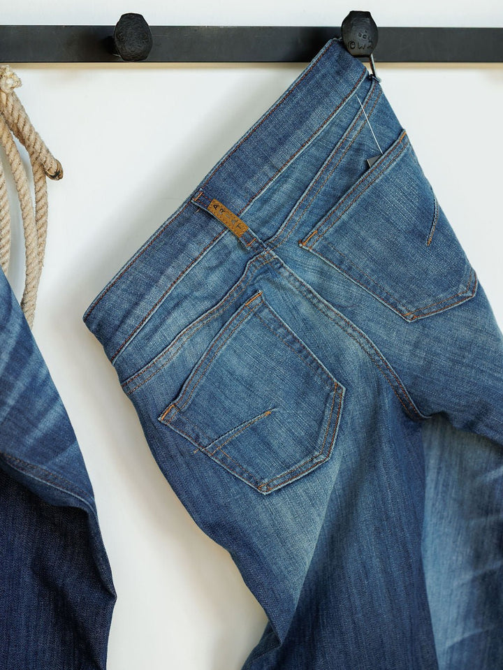 A pair of Ariat The Payson Perfect Rise Slim Trouser Jeans in medium dark wash hang by a belt loop on a black wall hook, showing back pockets and faded details, with a coiled rope beside them.