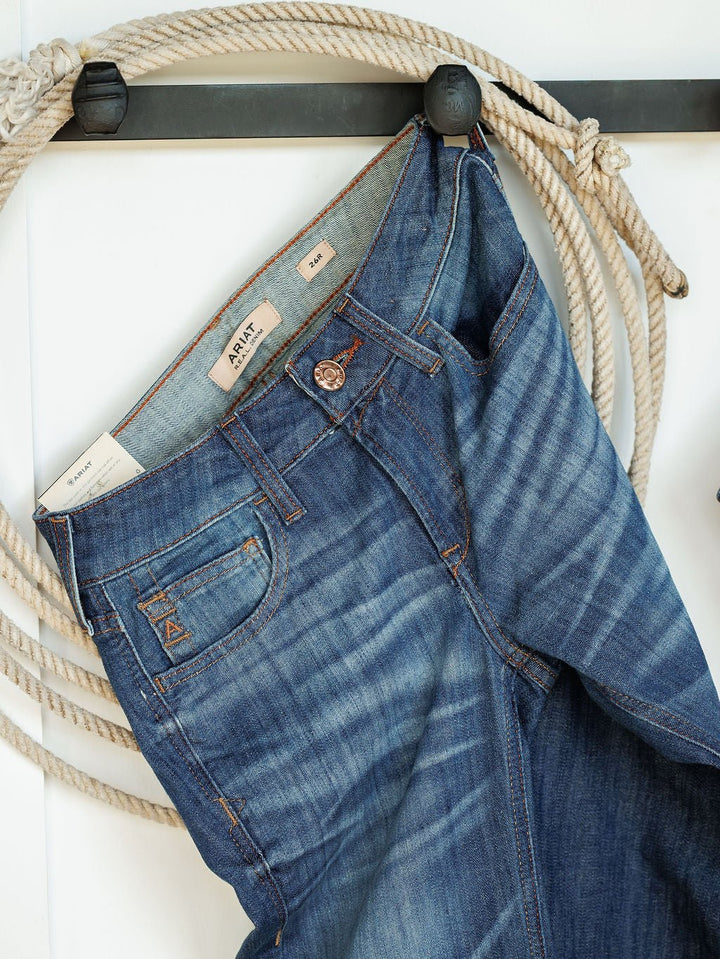 A pair of Ariat The Payson Perfect Rise Slim Trouser Jeans in medium dark wash hangs on a hook beside coiled rope, showing labels and pockets against a white backdrop.