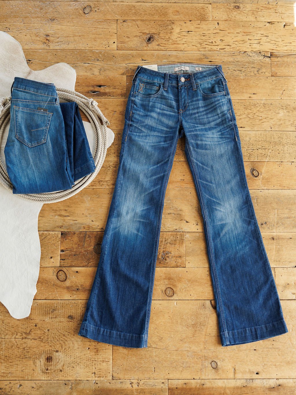 A pair of Ariat denim jeans is displayed flat on a wooden floor beside The Payson Perfect Rise Slim Trouser Jean, which is folded in a round rope-handle basket; part of a white animal hide appears on the left.