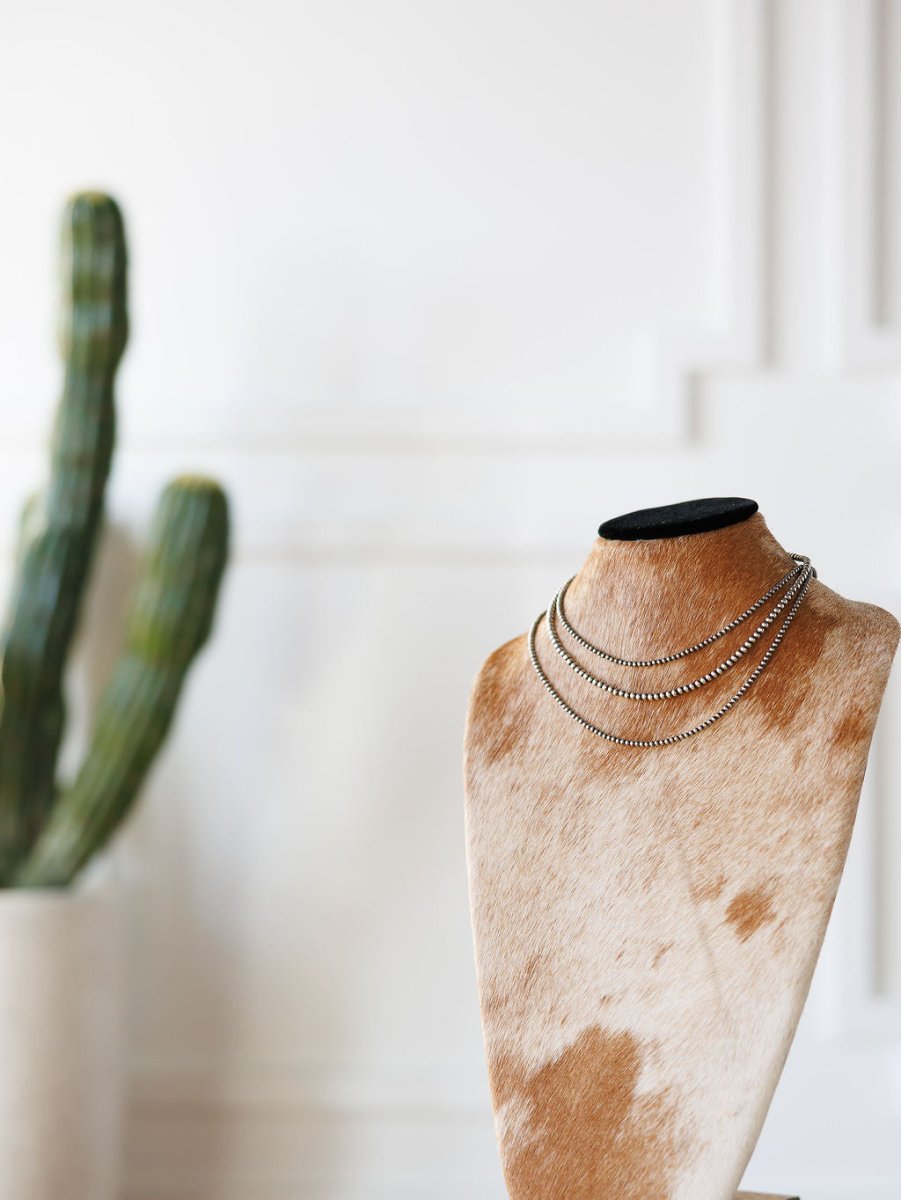 The Pecos Necklace by Savannah Sevens western life{&}style, a hand-strung sterling silver piece, is shown on a cowhide-covered jewelry bust with a green potted cactus blurred in the background against a white wall.