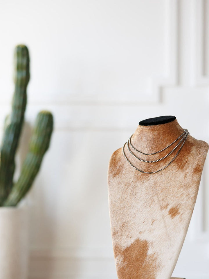 The Pecos Necklace by Savannah Sevens western life{&}style, a hand-strung sterling silver piece, is shown on a cowhide-covered jewelry bust with a green potted cactus blurred in the background against a white wall.