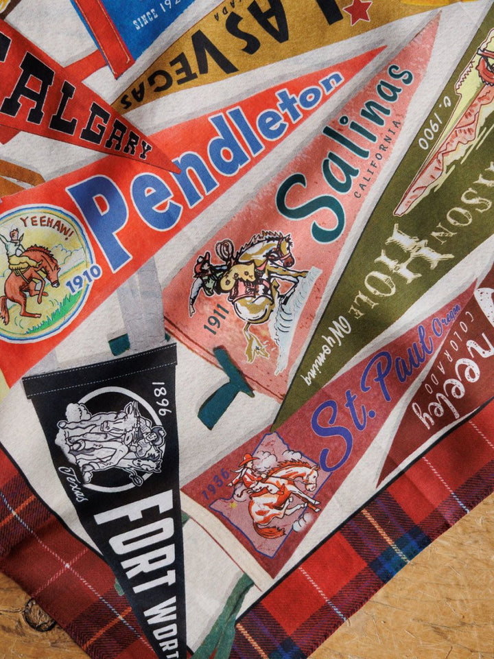 A detailed shot of the Pennants Neck Scarf by Savannah Sevens western life{&}style, featuring colorful vintage rodeo pennant designs with bucking broncos and city names, displayed on a plaid background.