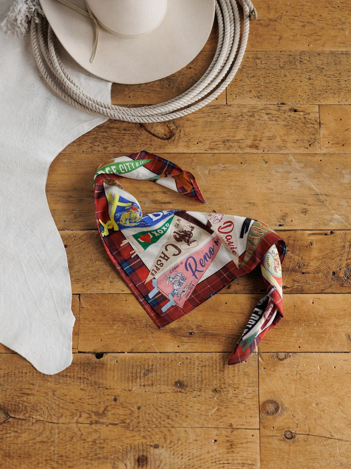 The Pennants Neck Scarf from Savannah Sevens western life{&}style, featuring a colorful pattern on cotton silk, is folded on a wooden floor next to a white cowboy hat and a piece of white, animal hide-like fabric.