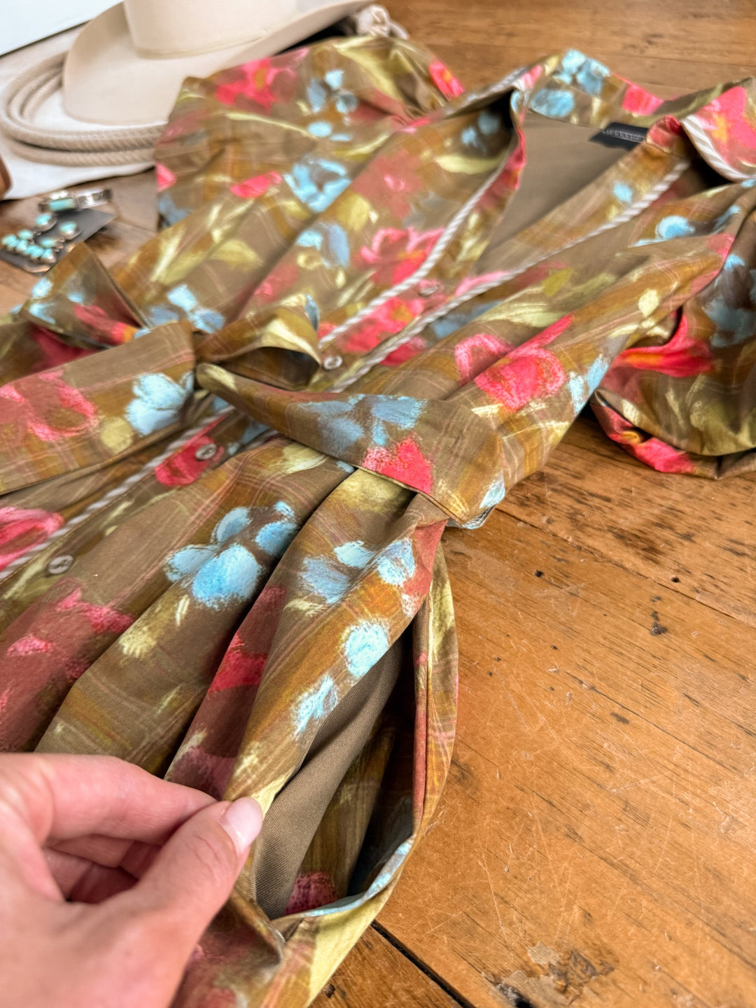 A hand pulls at the pocket of the Pensacola Olive Floral Midi Dress by Savannah Sevens western life{&}style—its red, blue, and green floral hues displayed on a wooden surface beside a hat and turquoise jewelry.