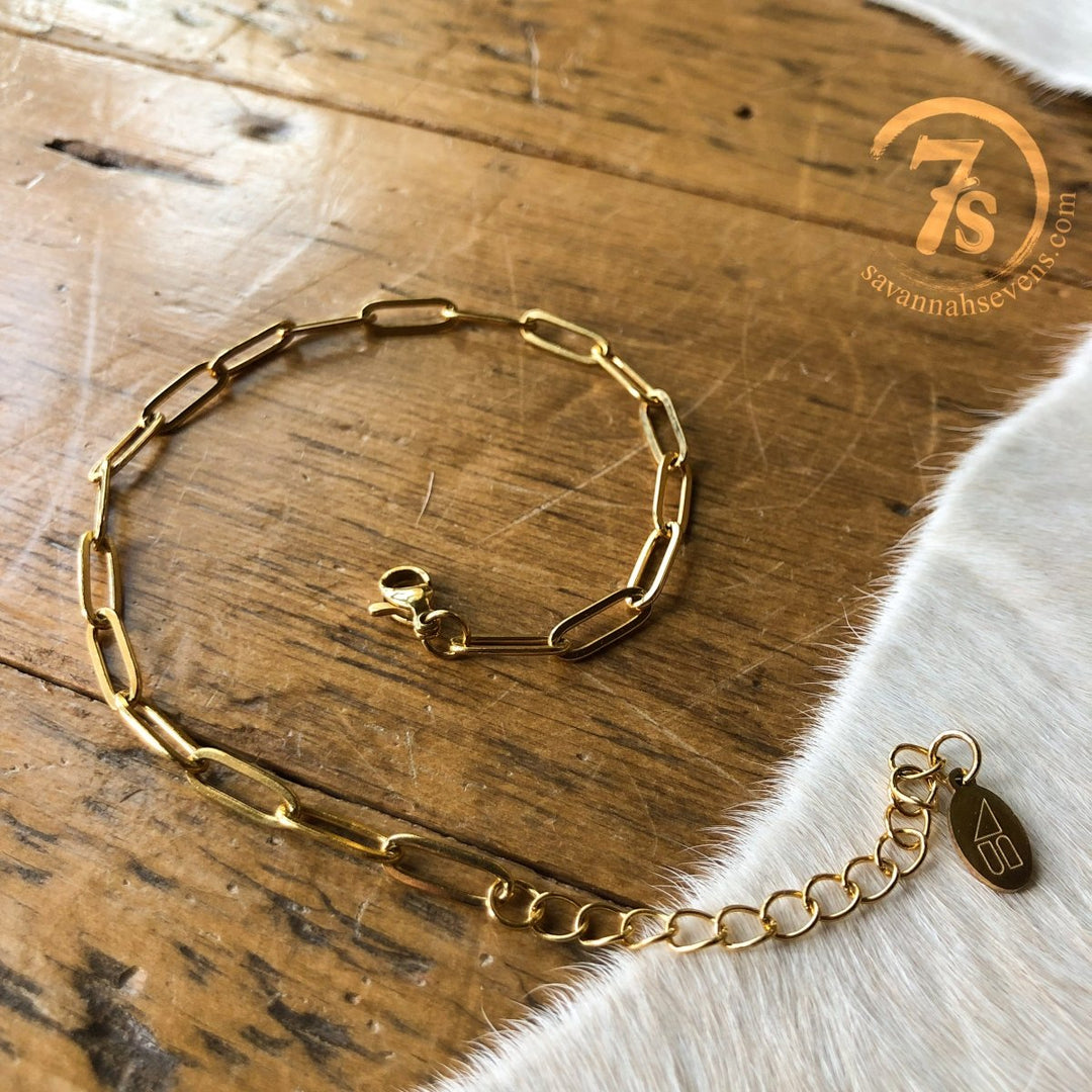 The Peralta Anklet by Savannah Sevens western life{&}style features elongated 14k gold filled links and a small tag, displayed on wood beside white fur. Tarnish resistant, it brings timeless elegance. 7s  logo in the top right corner.