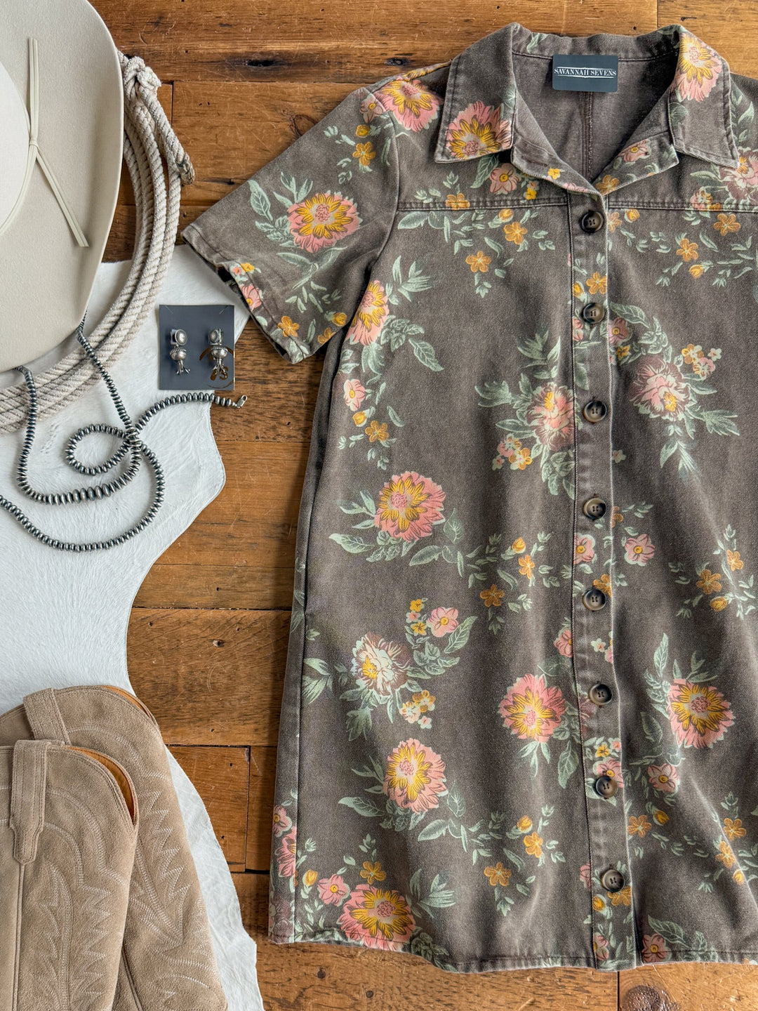The Petaluma Floral Button Up Dress by Savannah Sevens western life{&}style, with its short sleeves and cotton fabric, is styled on a wooden surface alongside a white cowboy hat, silver jewelry, and tan cowboy boots for a Western-inspired flat lay ensemble.