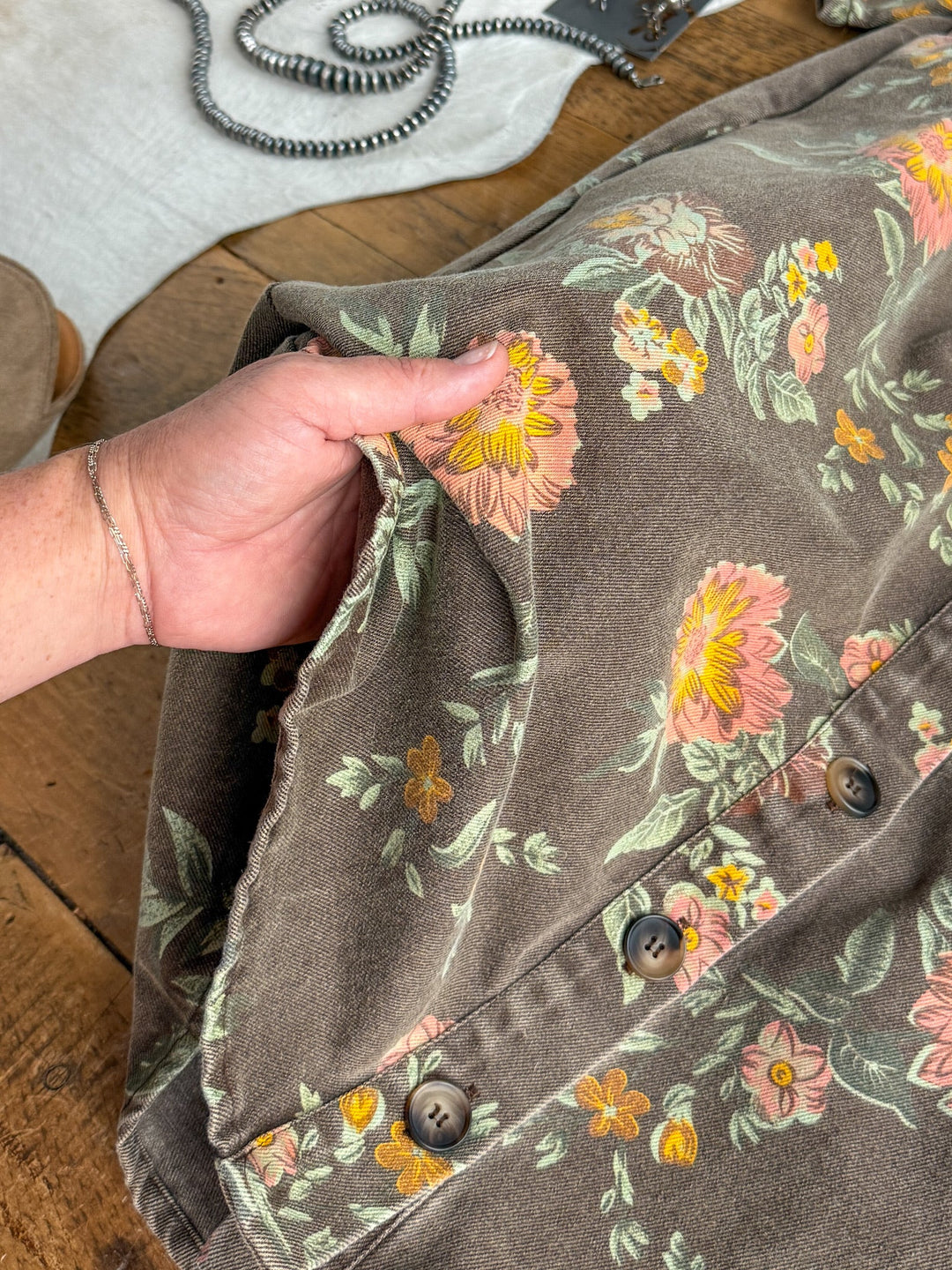 A hand holds the side pocket of a brown floral-patterned dress adorned with large pink and yellow flowers and green leaves, resting on a wooden surface beside a gray strap and part of a shoe—an inviting detail that perfectly complements the Petaluma Floral Button Up Dress from Savannah Sevens western life{&}style.