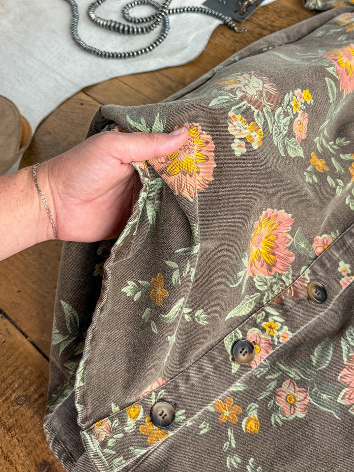 A hand holds the side pocket of a brown floral-patterned dress adorned with large pink and yellow flowers and green leaves, resting on a wooden surface beside a gray strap and part of a shoe—an inviting detail that perfectly complements the Petaluma Floral Button Up Dress from Savannah Sevens western life{&}style.