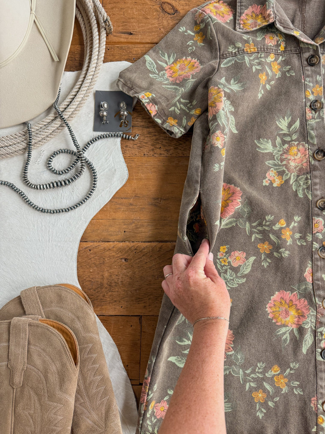 A hand shows the pocket of the Petaluma Floral Button Up Dress by Savannah Sevens western life{&}style on a wooden surface, surrounded by a hat, earrings, a necklace, a pair of boots, and a white cowhide rug.
