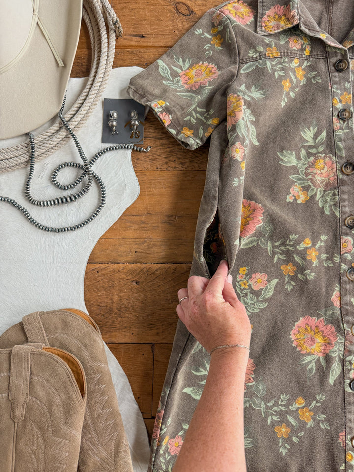 A hand shows the pocket of the Petaluma Floral Button Up Dress by Savannah Sevens western life{&}style on a wooden surface, surrounded by a hat, earrings, a necklace, a pair of boots, and a white cowhide rug.