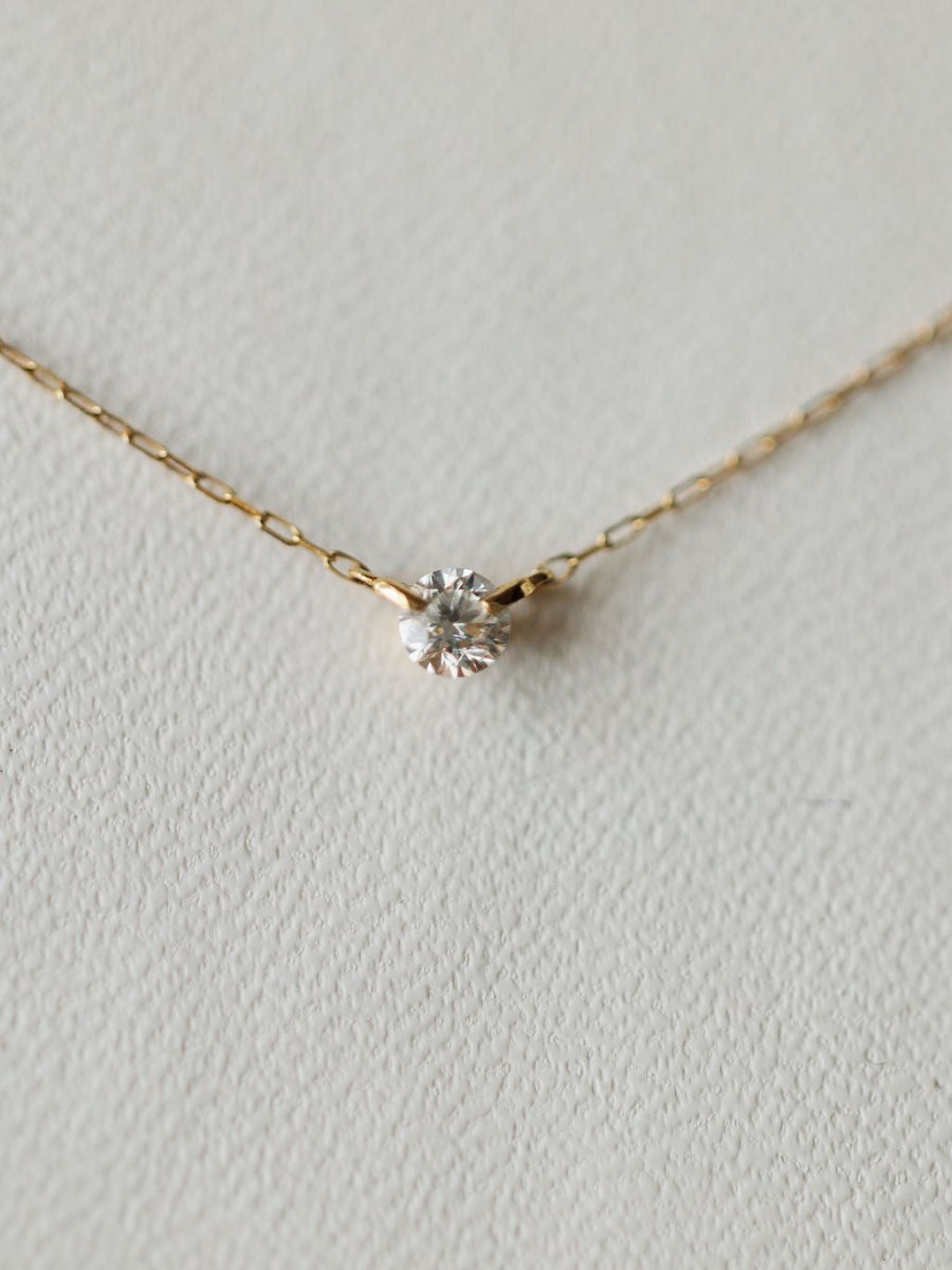 The Petite Diamond Necklace {Gold} by Savannah Sevens western life{&}style showcases a bezel set diamond pendant in 18K yellow gold, displayed on a light textured surface.