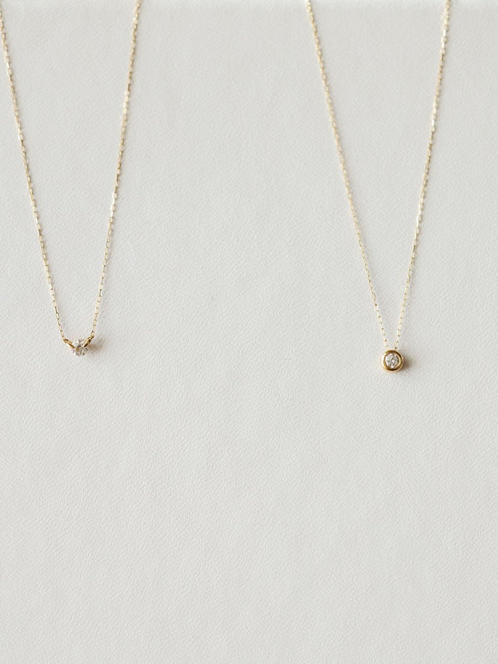 Two delicate gold necklaces by Savannah Sevens western life{&}style rest on a light, textured surface. The left is the Petite Diamond Necklace {Gold}; the right showcases a round, bezel-set diamond pendant.