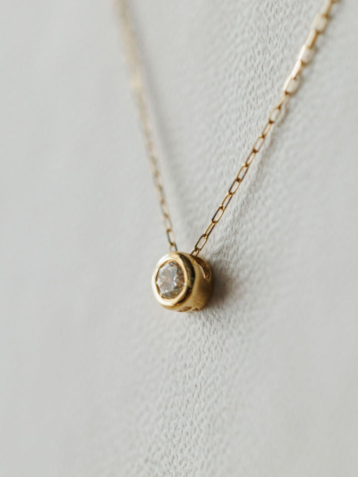 A close-up of the Petite Diamond Necklace {Gold} by Savannah Sevens western life{&}style, featuring a delicate 18K yellow gold chain and small bezel-set diamond pendant, displayed on a light textured background.