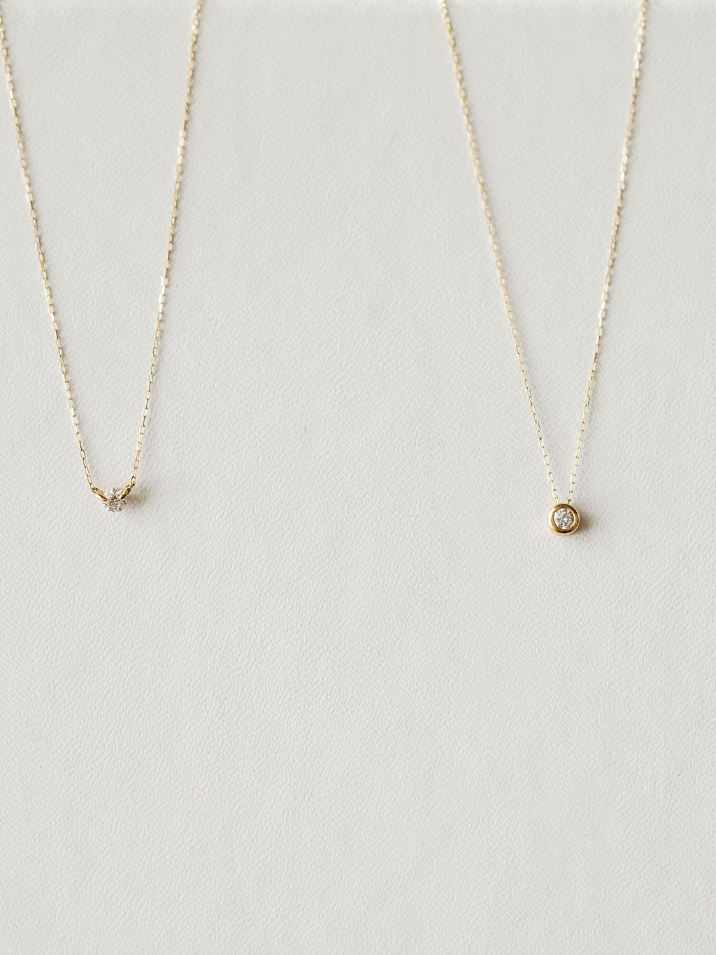Two delicate gold necklaces by Savannah Sevens western life{&}style rest on a light, textured surface. The left is the Petite Diamond Necklace {Gold}; the right showcases a round, bezel-set diamond pendant.