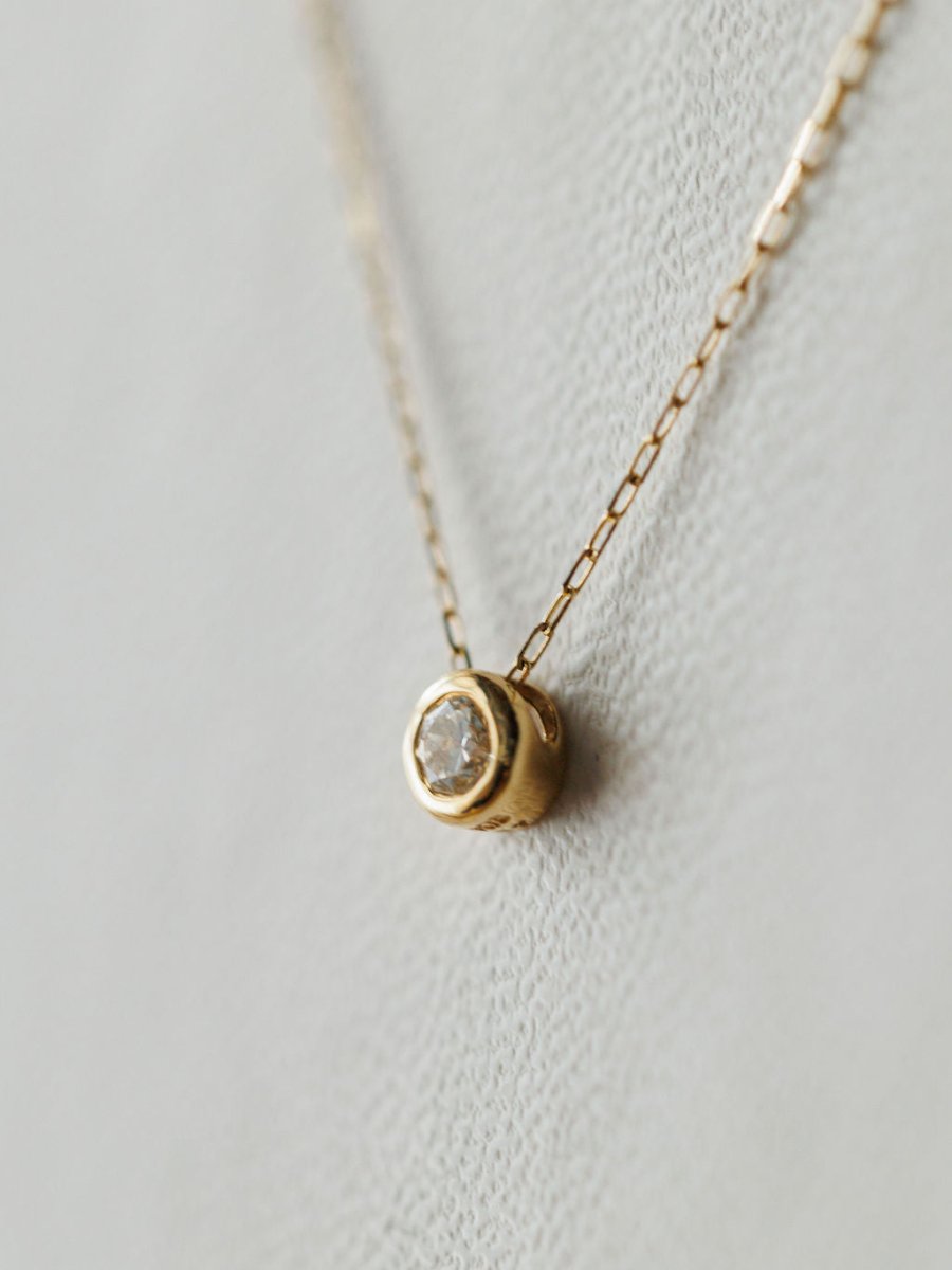 A close-up of the Petite Diamond Necklace {Gold} by Savannah Sevens western life{&}style, featuring a delicate 18K yellow gold chain and small bezel-set diamond pendant, displayed on a light textured background.