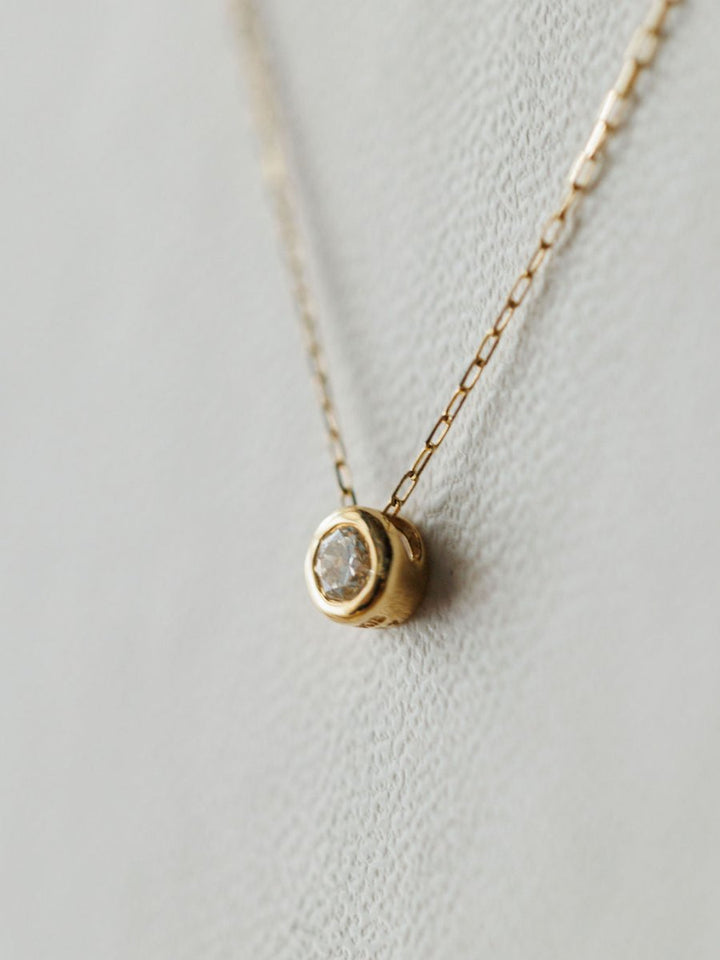 A close-up of the Petite Diamond Necklace {Gold} by Savannah Sevens western life{&}style, featuring a delicate 18K yellow gold chain and small bezel-set diamond pendant, displayed on a light textured background.