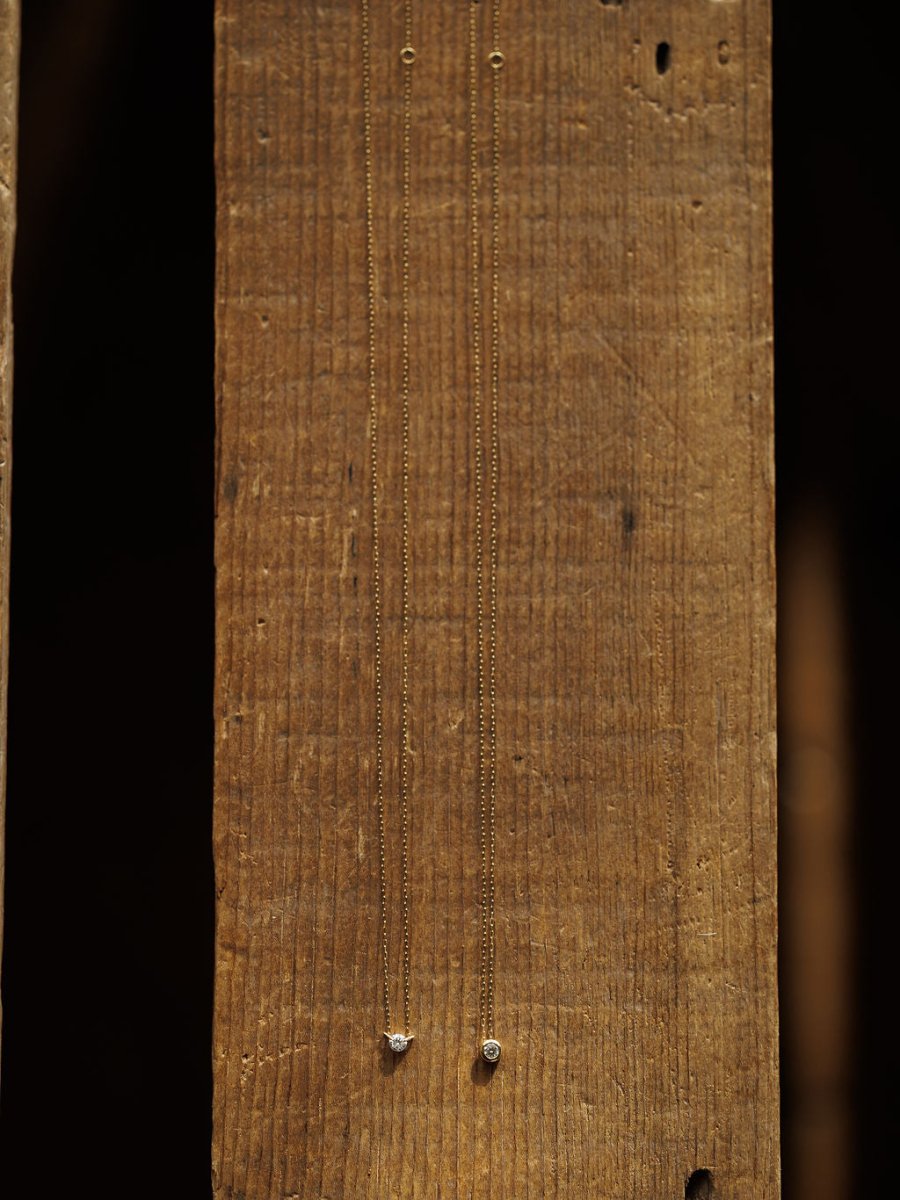 Two Petite Diamond Necklaces by Savannah Sevens western life{&}style, crafted in 18K yellow gold with small bezel-set diamonds, are displayed vertically against a rustic wooden background.