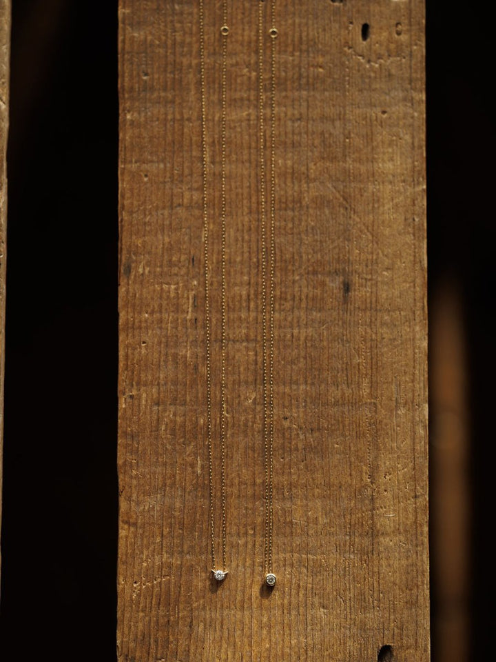 Two Petite Diamond Necklaces by Savannah Sevens western life{&}style, crafted in 18K yellow gold with small bezel-set diamonds, are displayed vertically against a rustic wooden background.