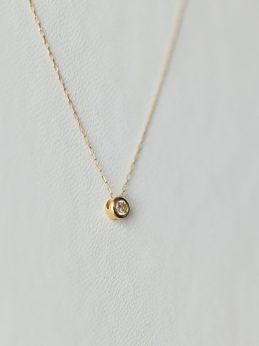 The Petite Diamond Necklace {Gold} by Savannah Sevens western life{&}style features a small round bezel-set diamond pendant on a delicate 18K yellow gold chain, shown against a light, textured background.