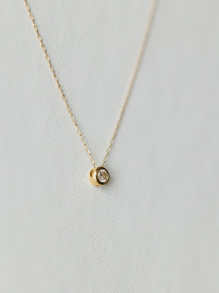 The Petite Diamond Necklace {Gold} by Savannah Sevens western life{&}style features a small round bezel-set diamond pendant on a delicate 18K yellow gold chain, shown against a light, textured background.