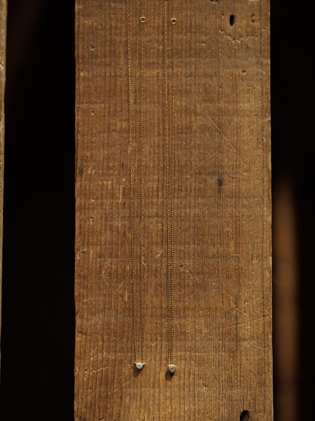 Two Petite Diamond Necklaces by Savannah Sevens western life{&}style, crafted in 18K yellow gold with small bezel-set diamonds, are displayed vertically against a rustic wooden background.