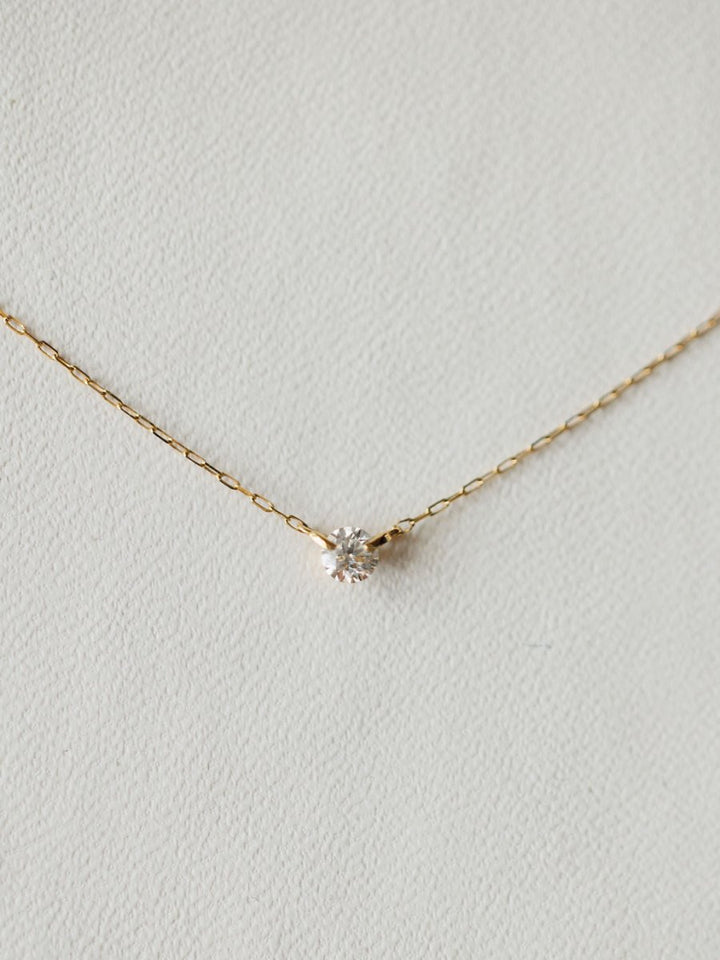 The Petite Diamond Necklace {Gold} by Savannah Sevens western life{&}style features an 18K yellow gold chain with a small, round bezel set diamond pendant, shown against a light, textured background.