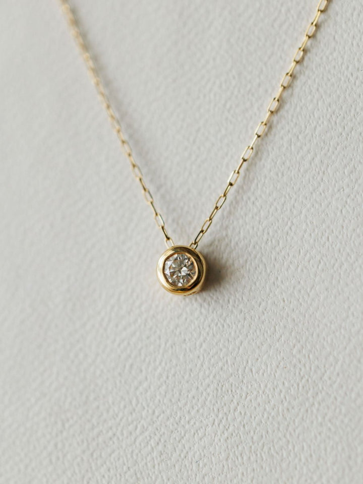 The Petite Diamond Necklace {Gold} by Savannah Sevens western life{&}style showcases a round bezel set diamond pendant in 18K yellow gold, elegantly displayed on a light textured background.