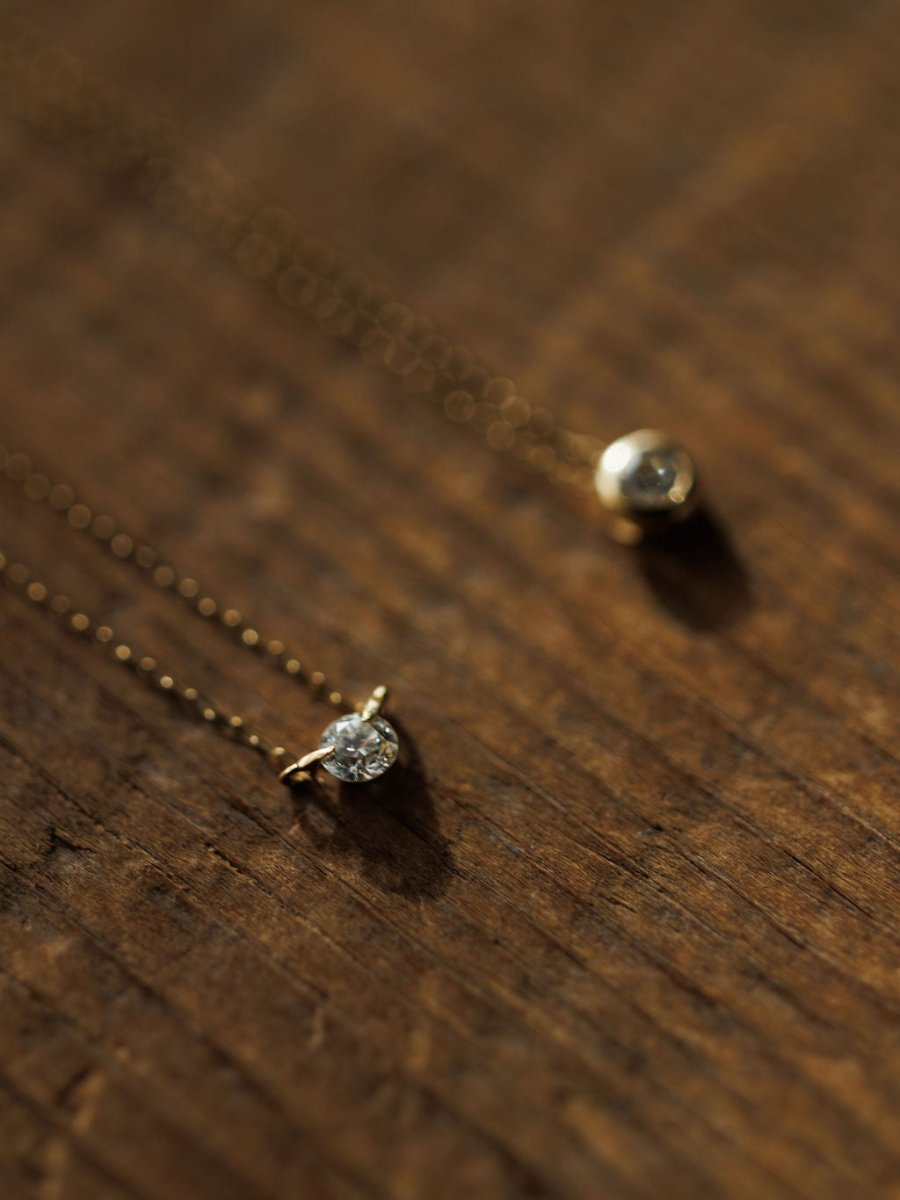 Two delicate Savannah Sevens western life{&}style Petite Diamond Necklace {Gold} pieces with small pendants—one a floating diamond, the other bezel set—rest on wood as warm light casts soft shadows.