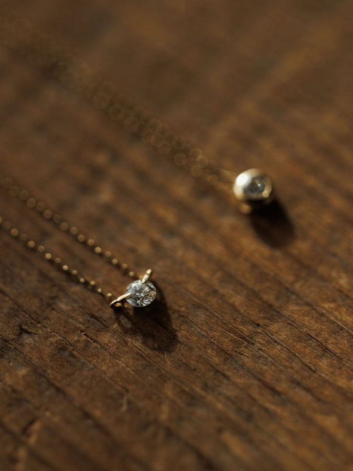 Two delicate Savannah Sevens western life{&}style Petite Diamond Necklace {Gold} pieces with small pendants—one a floating diamond, the other bezel set—rest on wood as warm light casts soft shadows.