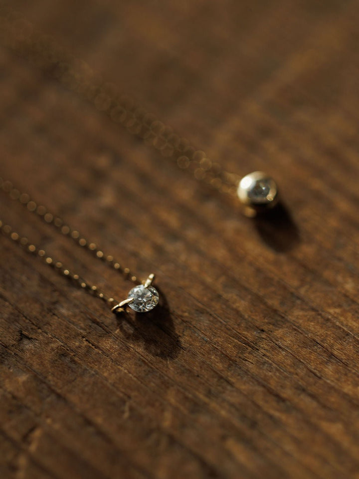 Two delicate Savannah Sevens western life{&}style Petite Diamond Necklace {Gold} pieces with small pendants—one a floating diamond, the other bezel set—rest on wood as warm light casts soft shadows.