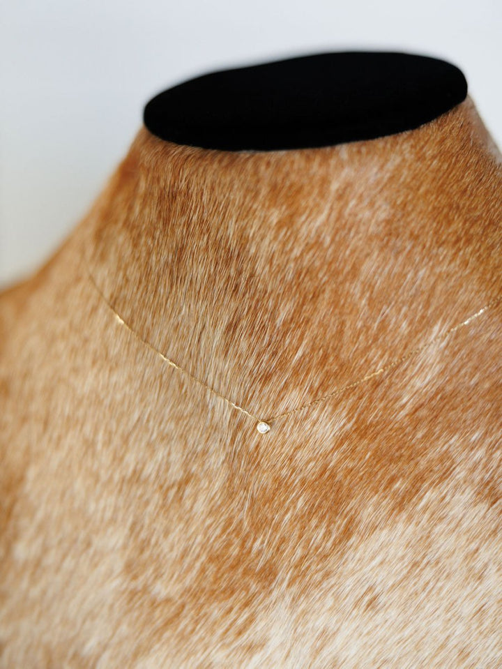 The Petite Diamond Necklace {Gold} by Savannah Sevens western life{&}style, featuring a small round bezel set diamond pendant, is displayed on a tan and white cowhide mannequin bust with a black top.