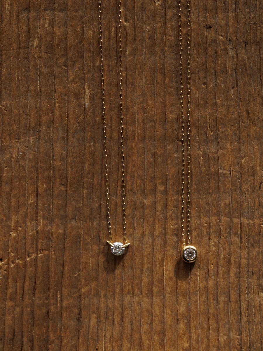 Two Petite Diamond Necklaces {Gold} by Savannah Sevens western life{&}style—one with a prong-set clear stone, the other with a bezel-set diamond—are displayed on wood with visible grain.