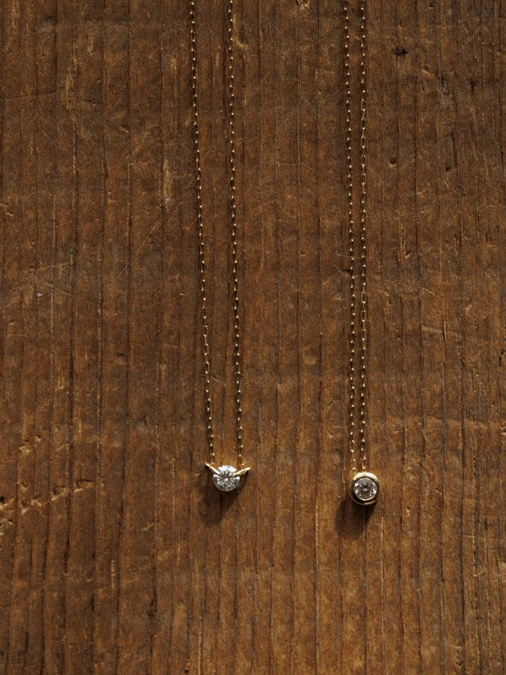 Two Petite Diamond Necklaces {Gold} by Savannah Sevens western life{&}style—one with a prong-set clear stone, the other with a bezel-set diamond—are displayed on wood with visible grain.