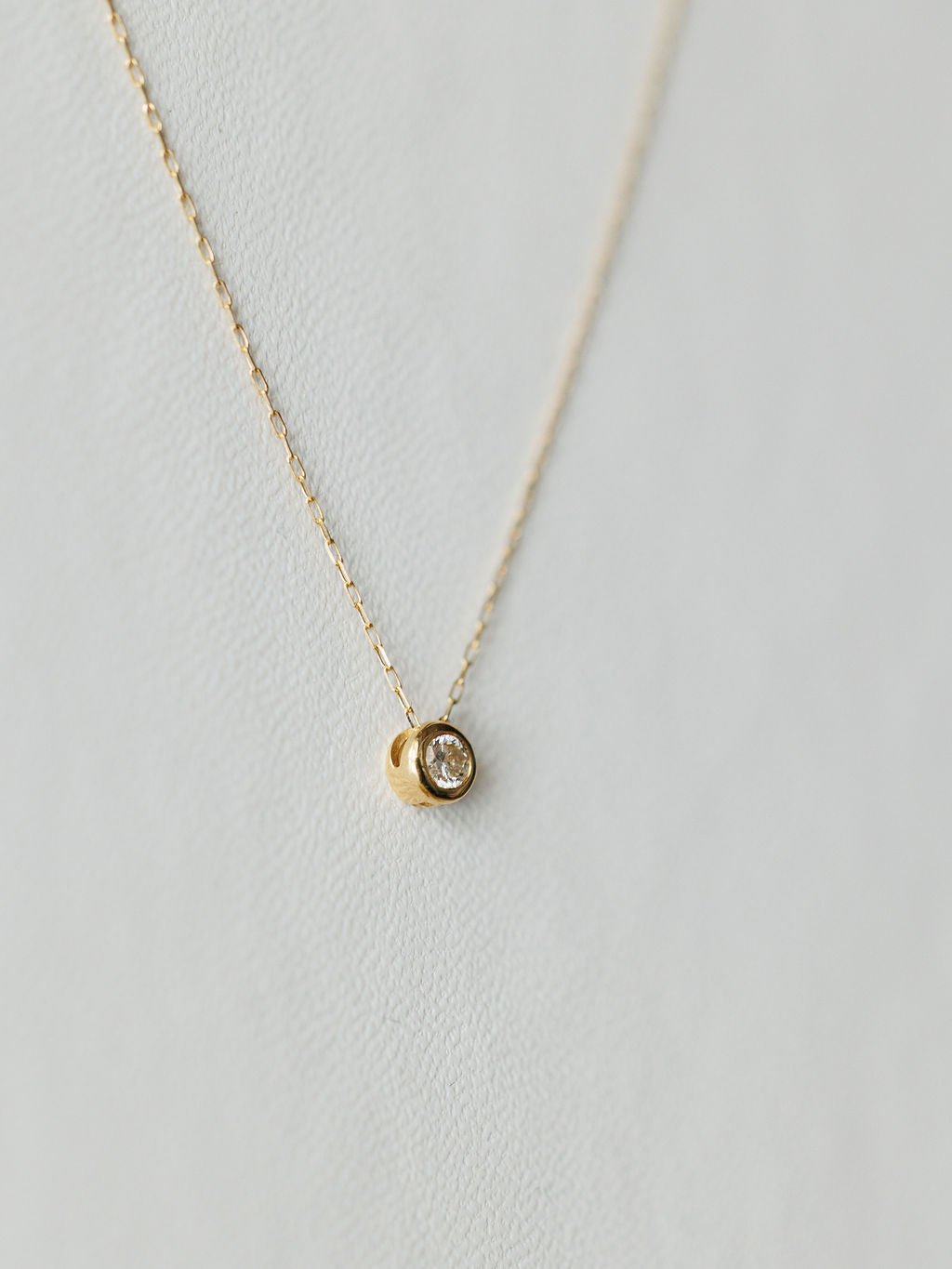 The Petite Diamond Necklace {Gold} by Savannah Sevens western life{&}style features a small round bezel-set diamond pendant on a delicate 18K yellow gold chain, shown against a light, textured background.
