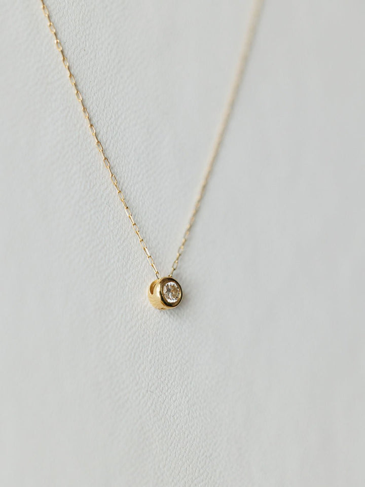 The Petite Diamond Necklace {Gold} by Savannah Sevens western life{&}style features a small round bezel-set diamond pendant on a delicate 18K yellow gold chain, shown against a light, textured background.