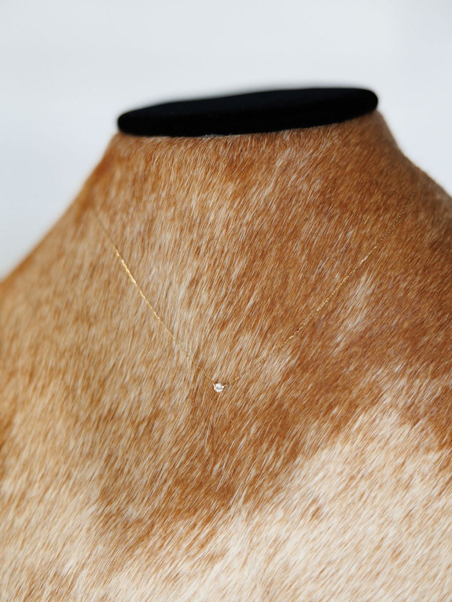 The Petite Diamond Necklace {Gold} by Savannah Sevens western life{&}style, featuring a bezel set diamond on an 18K yellow gold chain, is showcased on a brown and white cowhide mannequin bust against a plain background.