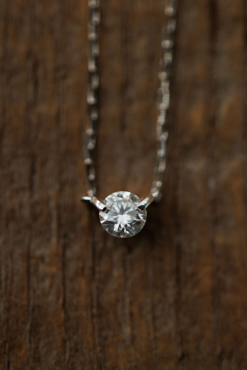 A close-up of the Petite Diamond Necklace {White Gold} by Savannah Sevens western life{&}style, featuring a natural round diamond pendant, displayed against a textured wooden background.