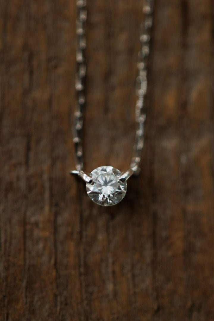 A close-up of the Petite Diamond Necklace {White Gold} by Savannah Sevens western life{&}style, featuring a natural round diamond pendant, displayed against a textured wooden background.