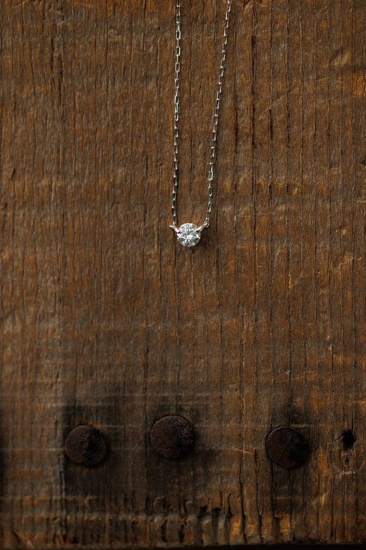 The Petite Diamond Necklace in White Gold by Savannah Sevens western life{&}style features a natural round diamond pendant, shining against a rustic wooden background.