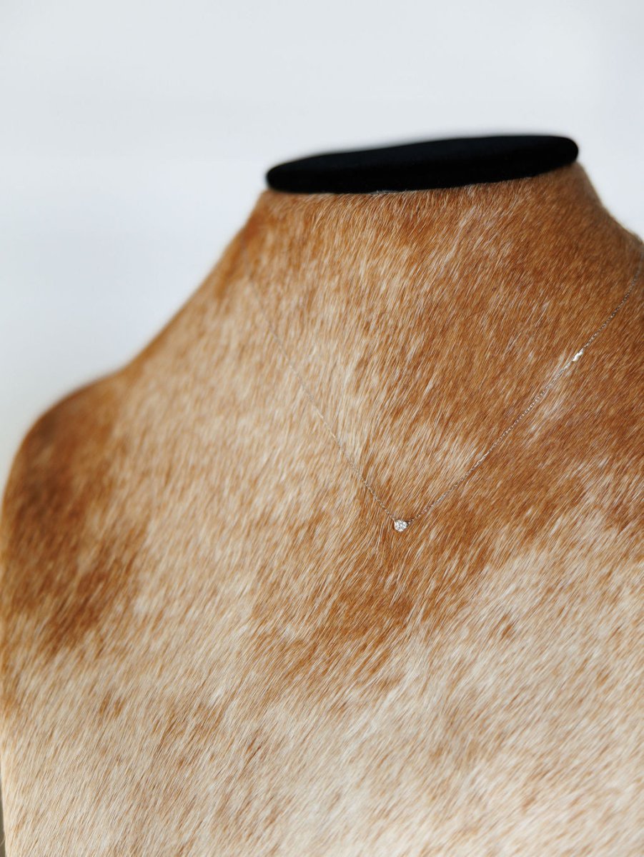 The Petite Diamond Necklace {White Gold} by Savannah Sevens western life{&}style, featuring a delicate solitaire pendant, is shown on a light brown and white cowhide mannequin against a simple, light background.