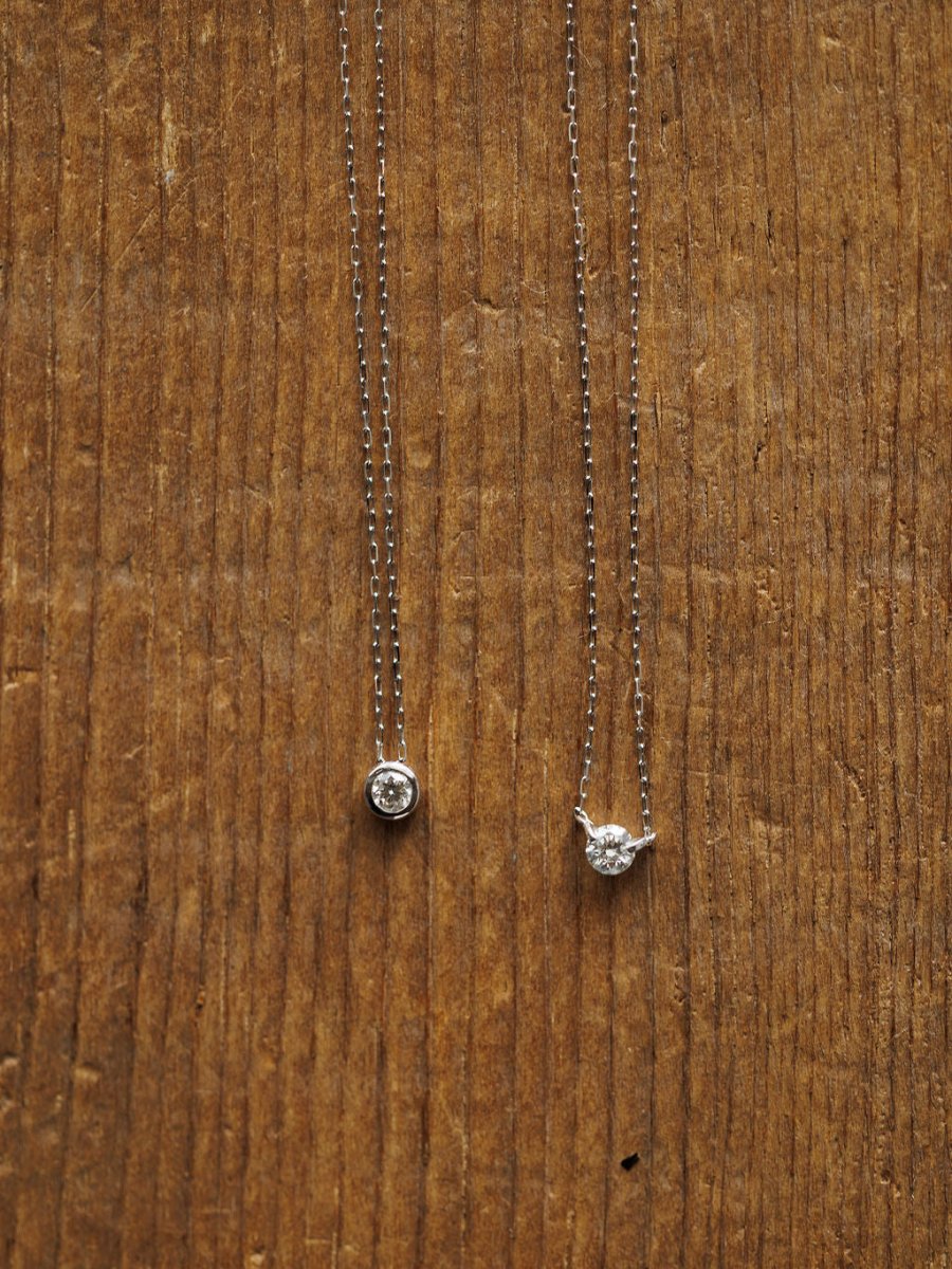 Two delicate Savannah Sevens western life{&}style necklaces are displayed: the Petite Diamond Necklace {White Gold} with a natural round diamond pendant and another featuring side prongs, both set on a textured wooden surface.