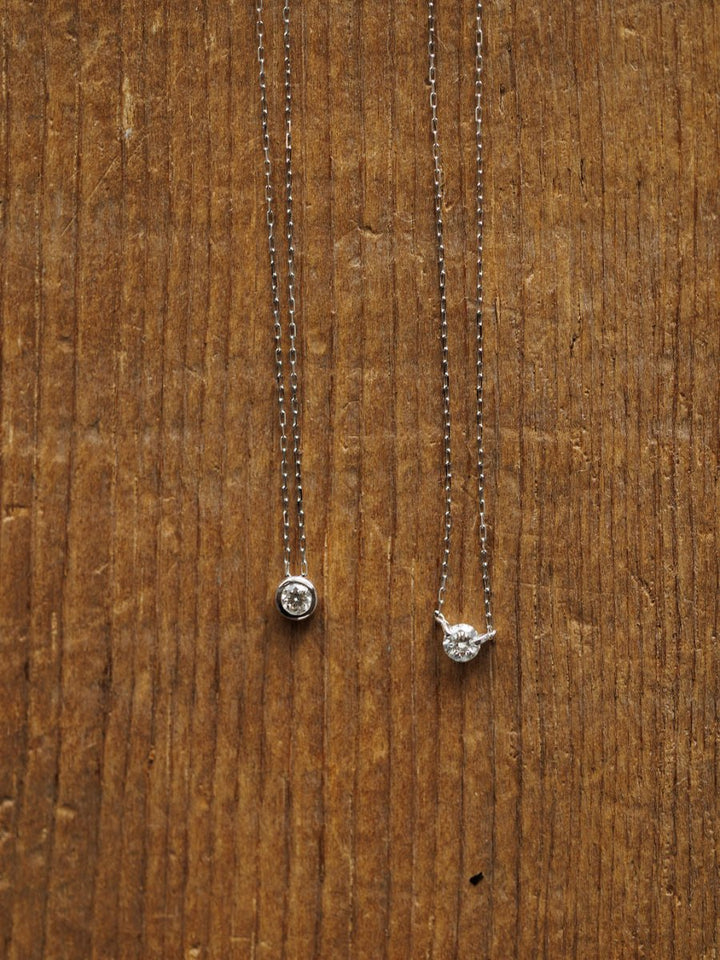 Two delicate Savannah Sevens western life{&}style necklaces are displayed: the Petite Diamond Necklace {White Gold} with a natural round diamond pendant and another featuring side prongs, both set on a textured wooden surface.