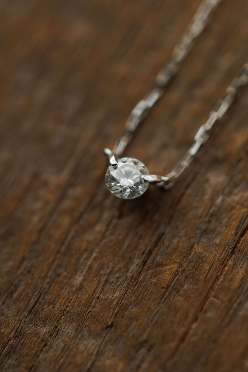 A close-up of the Savannah Sevens western life{&}style Petite Diamond Necklace {White Gold}, showcasing its natural round diamond pendant on a textured wooden surface.