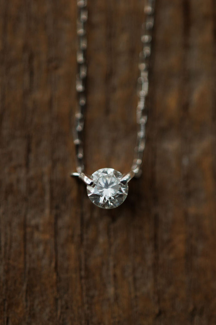 A close-up of the Petite Diamond Necklace {White Gold} by Savannah Sevens western life{&}style, featuring a natural round diamond pendant, displayed against a textured wooden background.