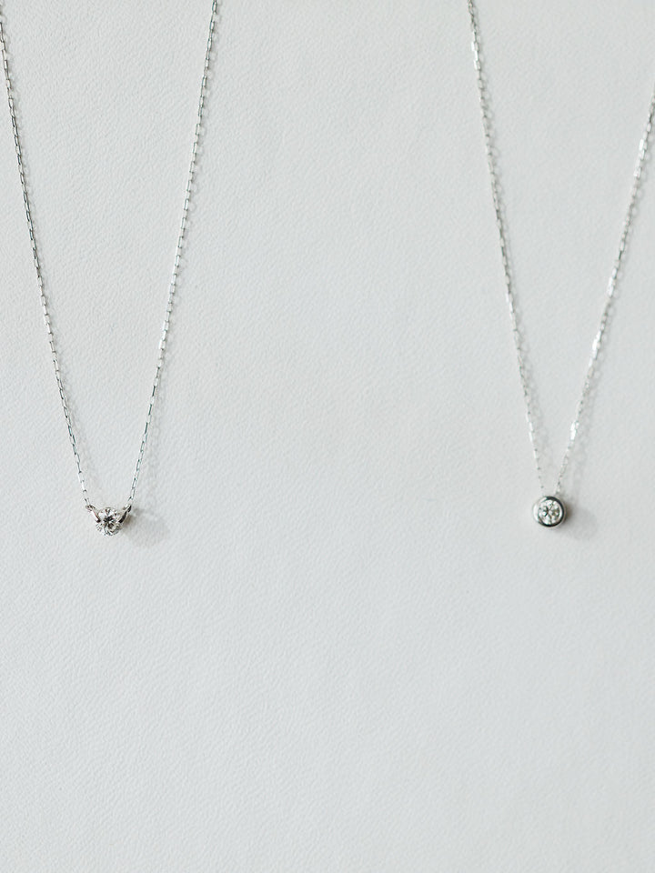 Two silver necklaces on a light background: the left has a small triangle pendant with stones, while the right is Savannah Sevens western life{&}style’s Petite Diamond Necklace in 18K white gold featuring a natural round diamond.